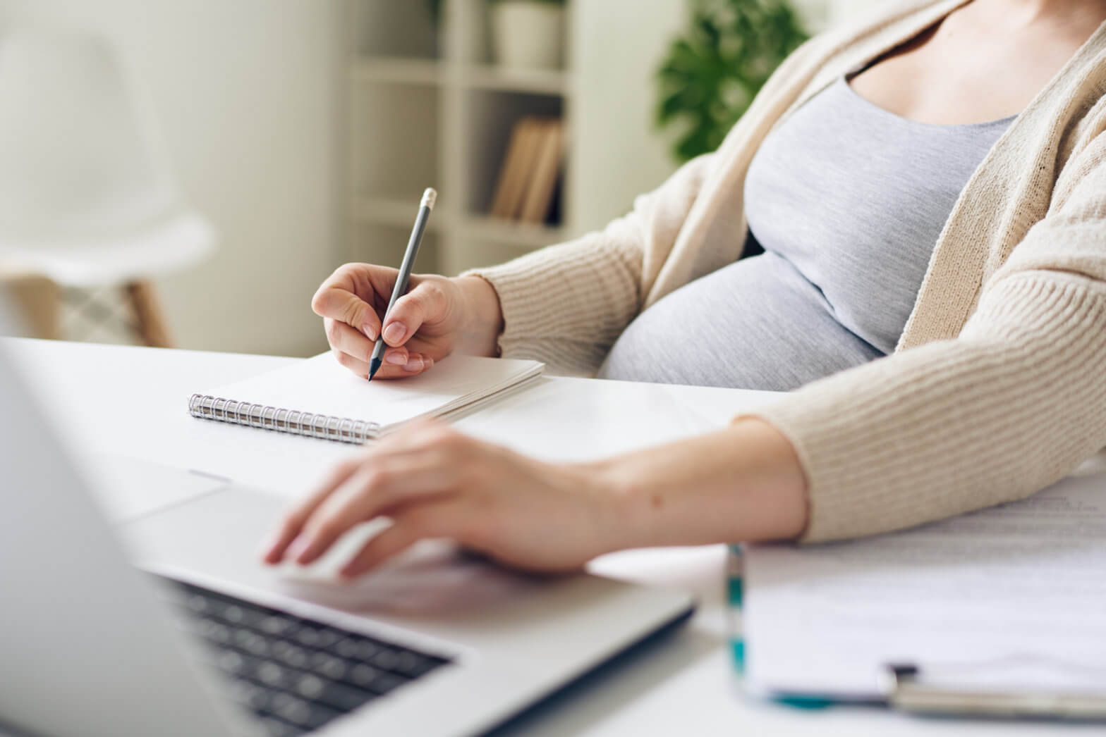 Close-up of unrecognizable pregnant woman sitting at table and using laptop while writing down tasks in notepad